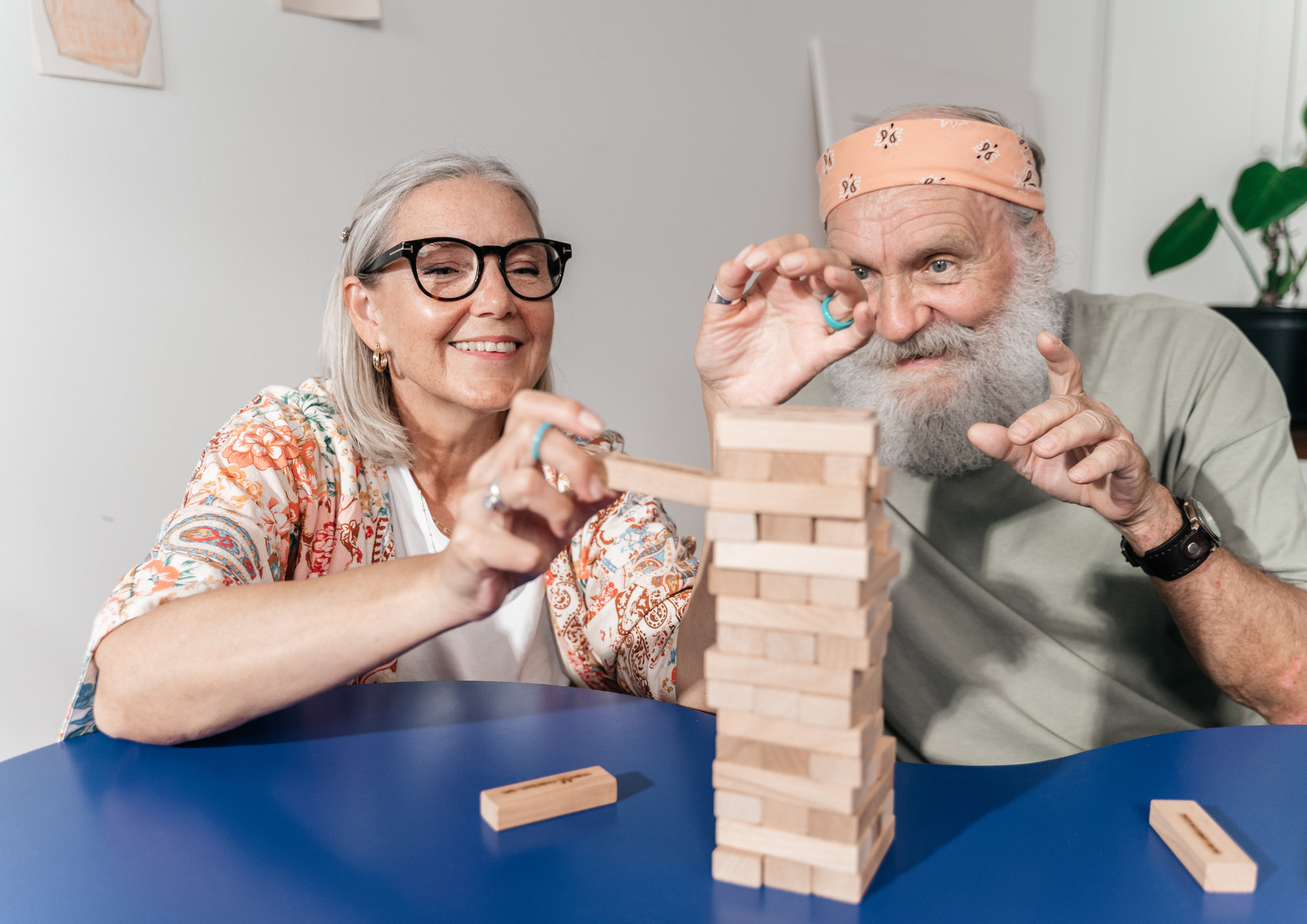 Seniorzy - mężczyzna i kobieta układają wieżę z klocków. Wyglądają na radosnych i zadowolonych. Kobieta ma czarne okulary w mocnych dużych oprawkach, a mężczyzna opaskę ze złożonej chustki na głowie.
