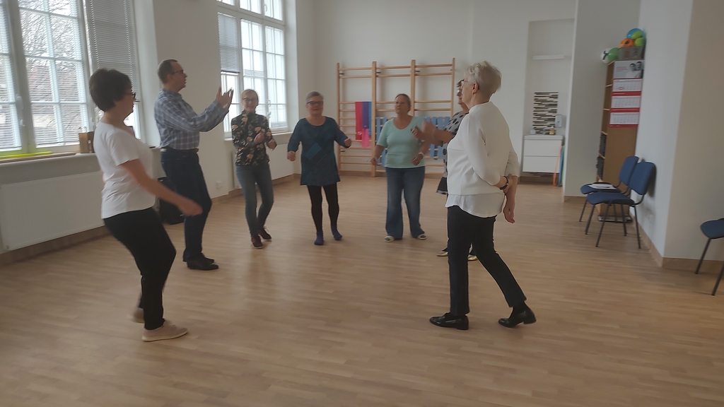 Dorosłe osoby stoją w pozycjach tanecznych. Mężczyzna, instruktor, coś im tłumaczy albo pokazuje za pomocą ustawienia ciała. Wszyscy znajdują się na sali gimnastycznej.