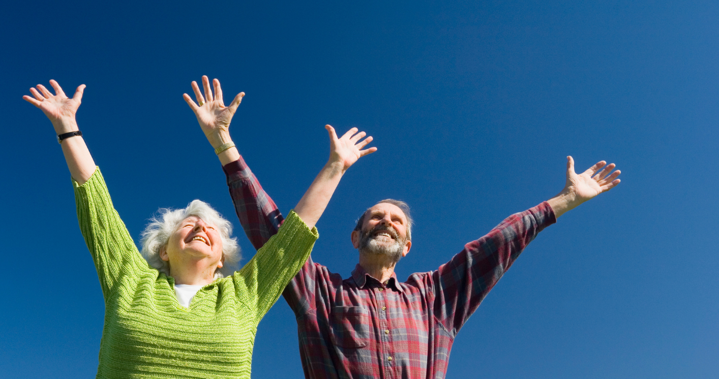 Na tle niebieskiego nieba stoją mężczyzna i kobieta w starszym wieku. Patrzą w górę, ich ręce są wysoko uniesione w geście radości.
