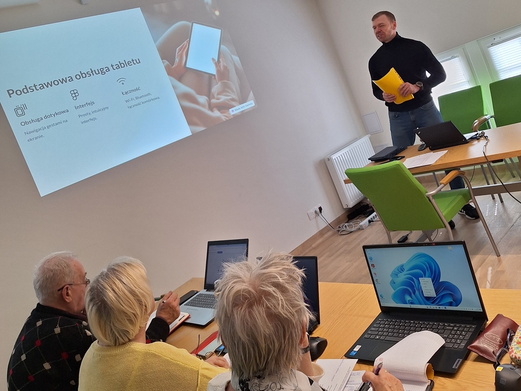 Sala komputerowa. Młody mężczyzna stojący przodem, trzyma w ręku kartki i coś tłumaczy osobom mobecnym na sali. Widać tylko głowy starszych osób. Od lewej, przy ścianie siedzi mężczyzna, a dalej w kierunku prawej strony dwie kobiet. Każde z nich siedzi przed uruchomionym laptopem. Na ścianie z lewej strony wyświetlony jest slajd.