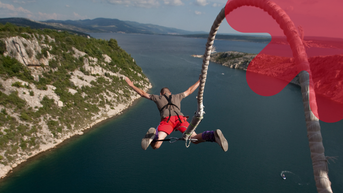 Mężczyzna w czerwonych szortach i uprzęży skacze na bungee z rozłożonymi szeroko rękami. Ujęcie od tyłu pokazuje skoczka zawieszonego nad rozległą zatoką otoczoną skalistymi, porośniętymi zielenią wzgórzami. Po prawej stronie widoczna jest gruba lina bungee, a w prawym górnym rogu – duży, półprzezroczysty czerwony symbol strzałki UniLodz.