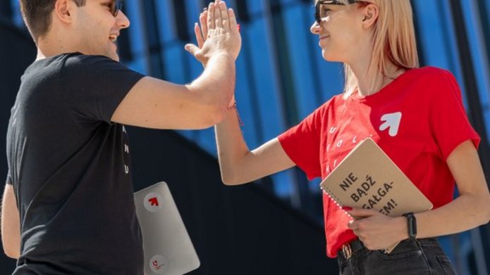 Dwoje młodych ludzi, dziewczyna i chłopak, stoją na dworze w słoneczny dzień i przybijają sobie piątkę