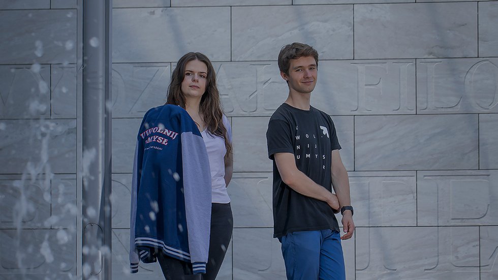 Studentka i student na tle szarej ściany z widocznymi wyrytymi literami. Na pierwszym planie tryskająca woda z fontanny.