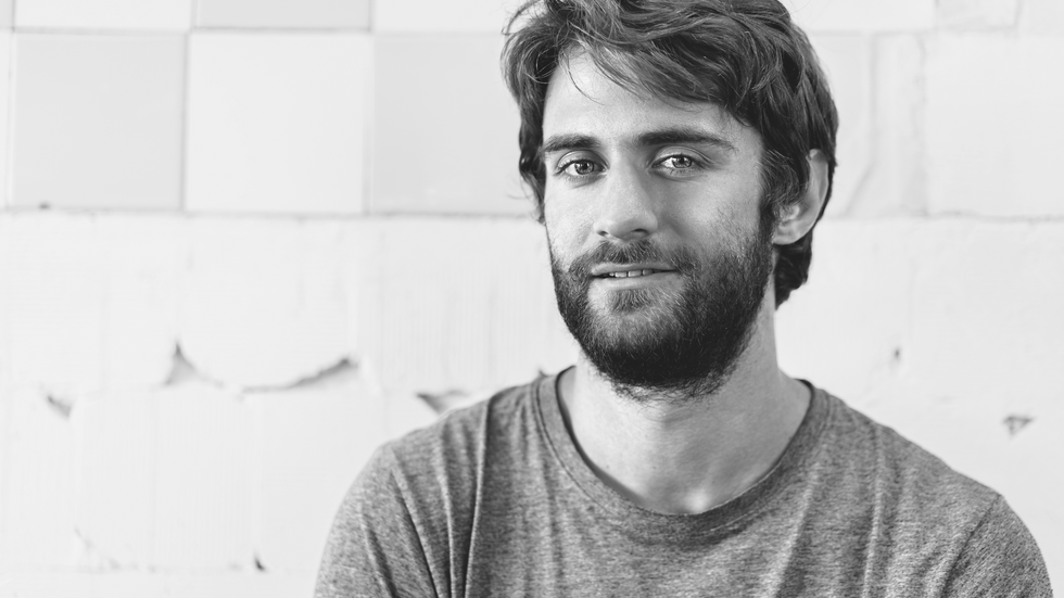 czarnobiałe zdjęcie przedstawiające uśmiechniętego młodego mężczyznę z brodą patrzącego wprost w obiektyw