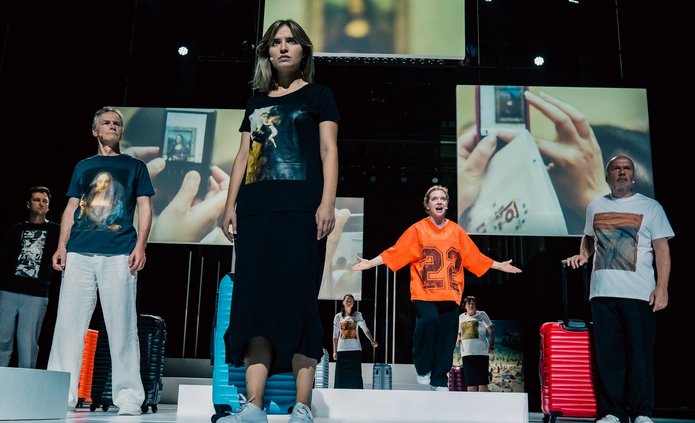 Na zdjęciu widoczna jest grupa osób stojących na scenie. Osoby są ubrane jak turyści, mają walizki. Za nimi wiszą różnej wielkości plansze na których są zdjęcia z różnych turystycznych miejsc.