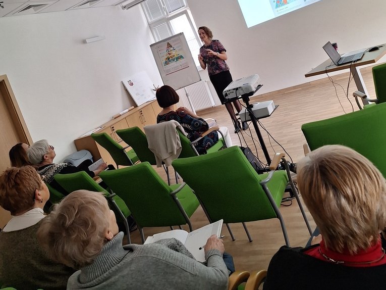 Sala wykładowa. Młoda kobieta stoi przed zasłuchaną publicznością. Na ekranie ściennym widocznym za jej plecami wyświetla się slajd przedstawiający przykład piramidy żywienia.